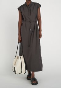 Person som bär en mörk ärmlös midiklänning med midjeband, bär en stor krämfärgad toteväska och svarta slidesandaler.