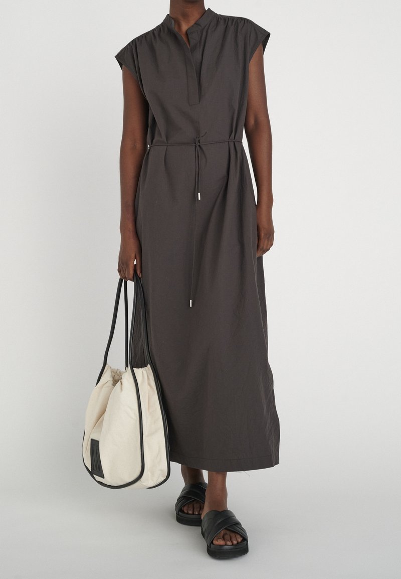 Person som bär en mörk ärmlös midiklänning med midjeband, bär en stor krämfärgad toteväska och svarta slidesandaler.