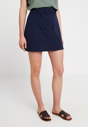 Mini-jupe bleu marine avec ceinture et détails de boutons, portée avec un top vert et des sandales tissées foncées sur une personne debout.