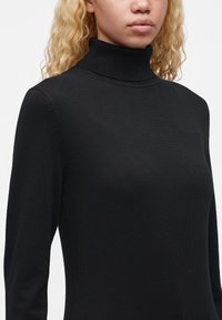 Femme aux cheveux blonds bouclés portant un pull noir ajusté à col roulé sur un fond clair uni.