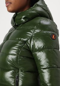 Groene pufferjack met een glanzende afwerking, gewatteerd ontwerp, capuchon en een oranje cirkelvormig merkembleem op de schouder. Voorzien van ritszakken.