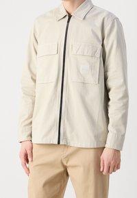 Jasno beżowa kurtka zapinana na zamek, wykonana z gładkiego materiału, z dwoma kieszeniami na piersi. Posiada czarny zamek i kwadratową naszywkę z logo na kieszeni.