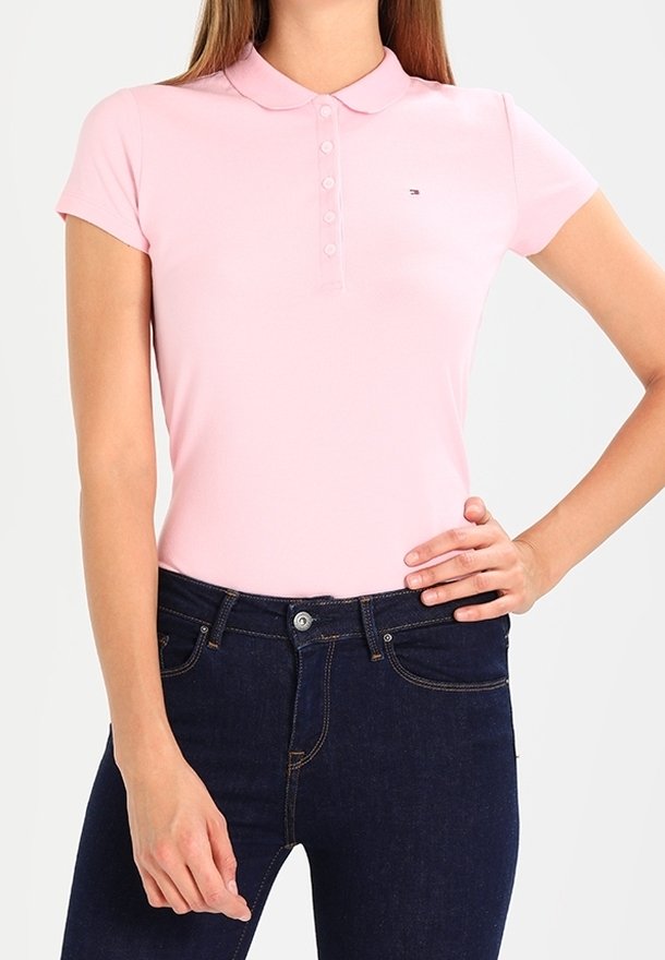 Femme portant un polo ajusté rose clair à manches courtes avec des boutons et un jean taille haute bleu foncé, main sur la hanche.