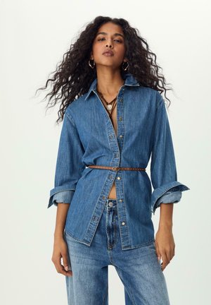 Jeanshemd in Mittelblau mit langen Ärmeln, durchgehendem Knopfverschluss und hochgekrempelten Manschetten. Kombiniert mit passenden Jeanshosen und einem braunen Ledergürtel.