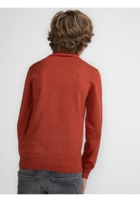 Maglione rosso lavorato a maglia con colletto alto, orlo a costine e maniche lunghe. La parte posteriore è liscia con una texture morbida, indossato con jeans grigi.
