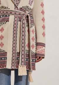 Cárdigan tejido de manga larga beige con patrones geométricos y florales en rojo, negro y azul, cinturón atado con flecos, llevado sobre jeans azules.