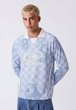 Jeune homme aux cheveux blonds courts portant une chemise à manches longues à carreaux bleus et blancs avec un col blanc et le logo "Heart in Lock".