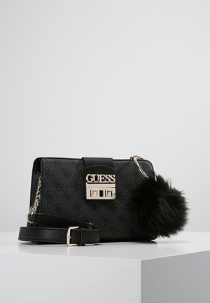 Sac bandoulière noir Guess avec fermoir logo, sangle amovible, détail chaîne et porte-clés pompon noir duveteux attaché sur une surface blanche.
