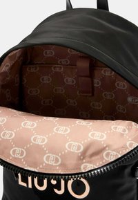 Zaino nero con apertura superiore con zip. L'interno presenta una fodera rosa con un motivo a logo ripetuto. Materiale liscio e forma strutturata.