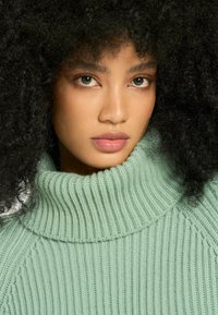 Grön ribbad polotröja med hög krage, med en mjuk textur och en åtsittande design, prydd med ett subtilt mönster längs ärmarna.