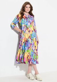 Abito midi floreale colorato con maniche a tre quarti, design a strati, dettaglio dei bottoni, in tonalità di blu, giallo e verde; abbinato a sabot bianchi.