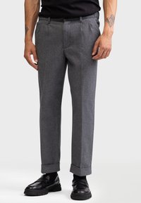 Pantalons en laine grise avec une coupe ajustée, présentant un motif chevrons discret, des plis sur les côtés et des ourlets retroussés, associés à des chaussures noires.