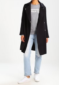 Zwarte lange jas over een grijze sweatshirt met witte tekst, blauwe versleten jeans en witte sneakers. De jas heeft een reverskraag en gouden knopen.