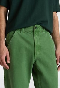 Green cotton pants with a straight cut, featuring a zip fly, belt loops, and side pockets. Texture appears slightly brushed.