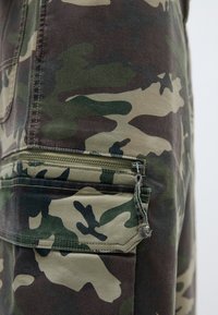 Kamuflāžas krāsas cargo bikses zaļā, brūnā un bēšā krāsā, ar sānu kabatu ar rāvējslēdzēju un izturīgu šuvēm, lai nodrošinātu ilgmūžību.