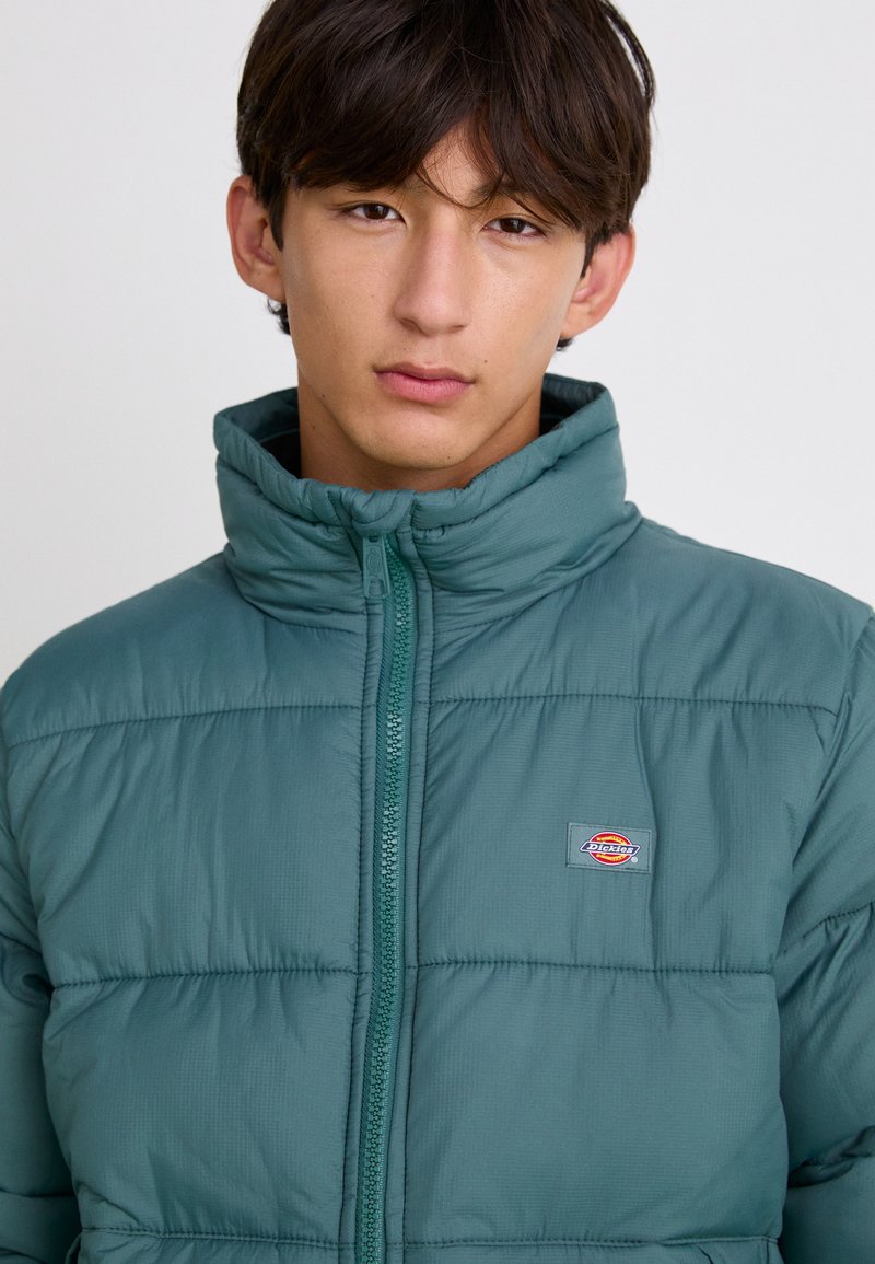 Junge Person in einer türkisfarbenen Pufferjacke mit Reißverschluss und kleinem Logo auf der Brust, die direkt in die Kamera blickt, vor einem einfarbigen Hintergrund.