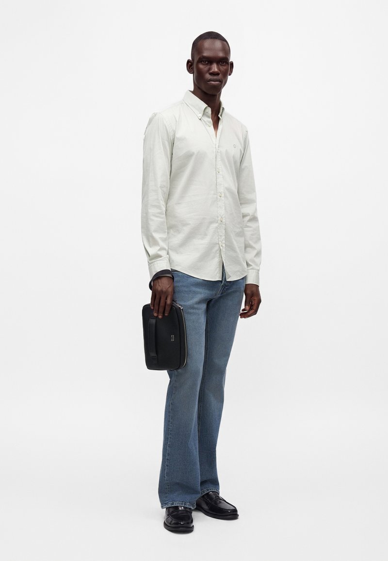 Homme portant une chemise blanche boutonnée, un jean bleu, des chaussures noires, et tenant une pochette rectangulaire noire avec une lanière au poignet.