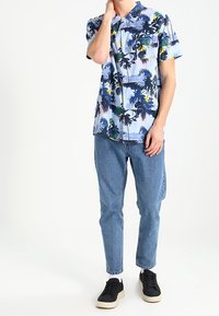 Kurzärmliges, knöpfbares Hemd mit blauem Hintergrund und tropischem Pflanzenmuster in Grün, Pink und Gelb. Getragen mit hellblauen Jeans und schwarzen Turnschuhen.