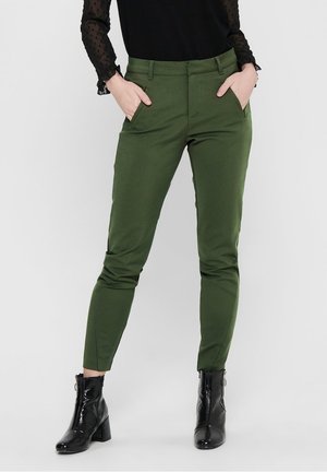 Kvinde iført tætsiddende olivengrønne bukser og sorte ankelstøvler med hæl, med hænderne i lommerne mod en ensfarvet baggrund.