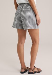 Donna che indossa degli shorts a righe verticali bianche e nere e sandali con cinturini marroni, in piedi su uno sfondo neutro chiaro.