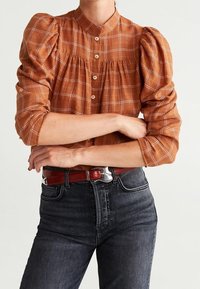 Blouse à carreaux bruns avec des manches bouffantes, devant boutonné et plis. Associée à un jean en denim noir et une ceinture rouge. Tissu texturé.