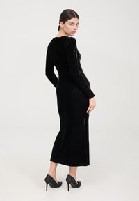 Abito midi in velluto nero con maniche lunghe, silhouette aderente e scollatura a U. Indossato con tacchi alti neri texture.