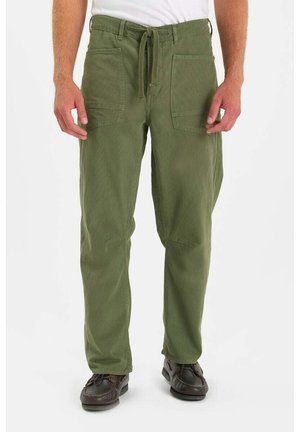 Man in olijfgroene broek met trekkoord en grote voorzakken, met donkerbruine schoenen, staand tegen een witte achtergrond.