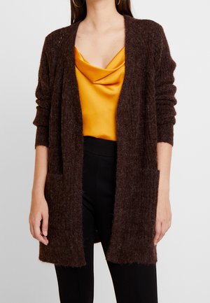 Frau trägt eine strukturierte dunkelbraune offene Strickjacke über einem senfgelben Satin-Top mit Wasserfallausschnitt und schwarzen Hosen.