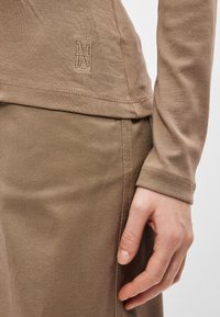 Κοντινό πλάνο ενός χεριού πο�υ ξεκουράζεται στο πλάι, φορώντας ένα μπεζ μακρύ μανίκι και ταιριαστά υφασμάτινα καφέ παντελόνια με κεντημένο λογότυπο.