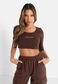 Brun cropped top med rund halsudskæring og lange ærmer, featuring et guld logo. Sammen med matchende brune bukser med elastisk talje.