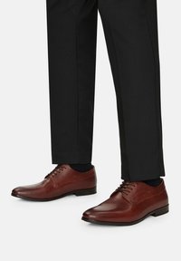 Hnědé kožené boty s hladkým povrchem, kulatou špičkou a tenkými tkaničkami, spárované s černými kalhotami a ponožkami. Mají nízký podpatek.