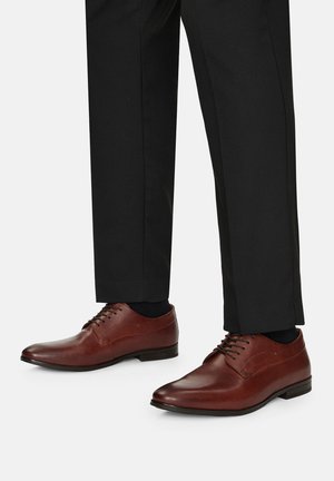 Hnedé kožené topánky s hladkým povrchom, okrúhlym špičkou a tenkými šnúrkami, v kombinácii s čiernymi nohavicami a ponožkami. Majú nízky opätok.