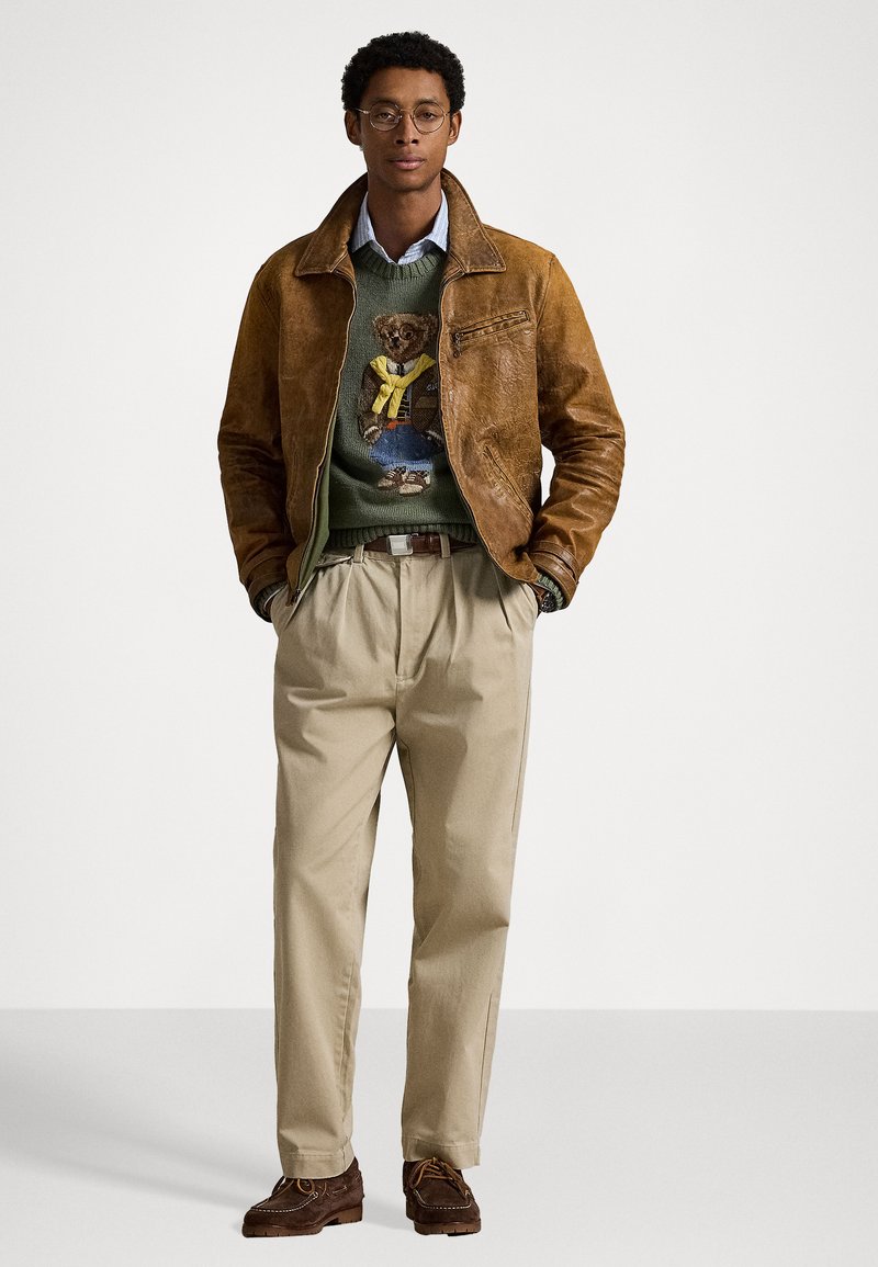 Giacca di pelle marrone, maglione verde con design di orso, pantaloni beige plissettati e scarpe marroni. Il modello è in piedi contro uno sfondo semplice.