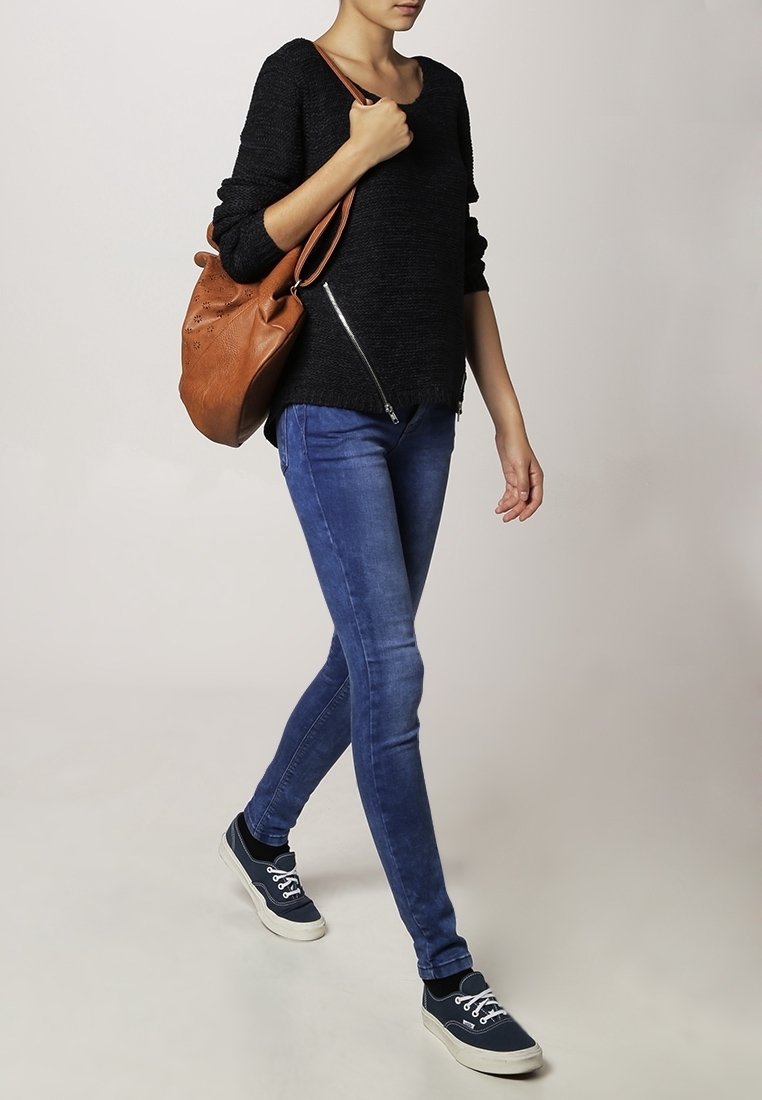 Femme marchant portant un jean bleu, un pull noir avec des fermetures éclair, des baskets noires, et portant un sac à bandoulière en cuir marron.