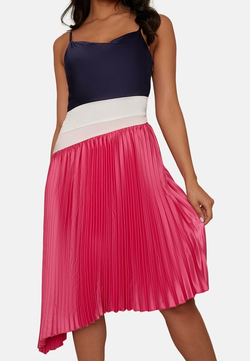 Faltender Rock aus pinkem Satin, mit einem marineblauen Oberteil und weißem Taillenbund. Der asymmetrische Saum verleiht eine dynamische Form. Insgesamt glatte Textur.