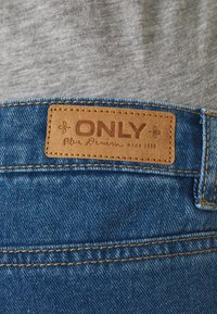 Jeans de ganga azuis com uma etiqueta de pele castanha rotulada "ONLY" e "Blue Denim anno 1995." Costura visível em fio dourado.