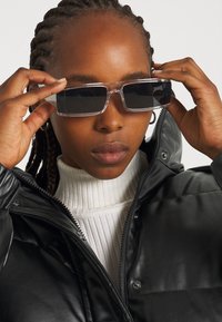 Gafas de sol con montura transparente y lentes oscuros. El modelo lleva una chaqueta acolchada negra y un cuello de tortuga blanco de canalé. Pelo trenzado.