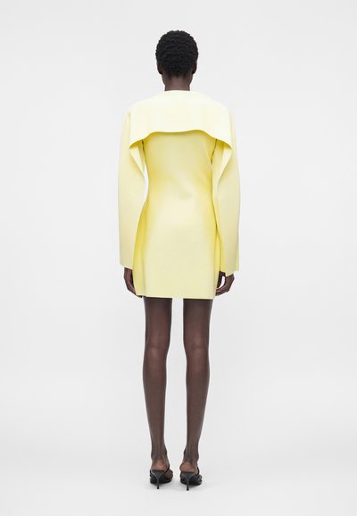 Γυναίκα με κοντά σγουρά μαλλιά που φοράει ένα εφαρμοστό μίνι φόρεμα σε ανοιχτό κίτρινο χρώμα με μακριά μανίκια και φαρδιά γιακά και μαύρα τακούνια σανδάλια, να στέκεται με την πλάτη.