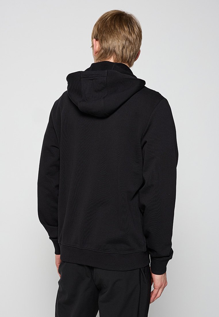 Mann mit kurzem hellbraunem Haar, der einen schwarzen Hoodie und schwarze Hosen trägt und mit dem Rücken zu einem schlichten hellgrauen Hintergrund steht.