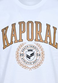 La camiseta blanca presenta un gran texto "KAPORAL" en marrón, con un gráfico circular que incluye "SOUTH VARSITY" y un escudo, rodeado de un diseño de laureles.