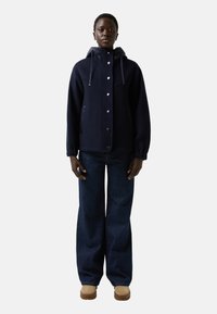 Granatowa kurtka z kapturem, z zatrzaskami i bocznymi kieszeniami, połączona z szerokimi ciemnoniebieskimi dżinsami i beżowym obuwiem.