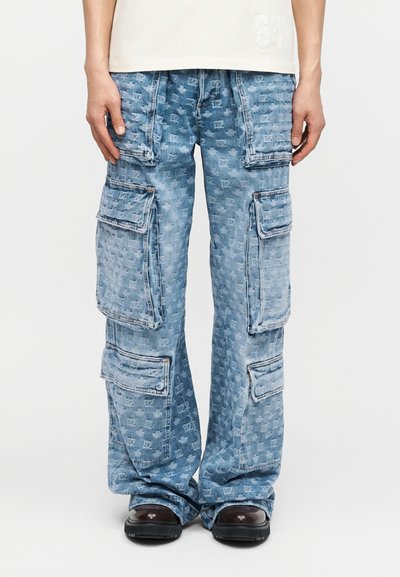 Pantalon cargo en denim bleu clair avec motif monogramme intégral et plusieurs grandes poches à rabat, porté avec des chaussures foncées et un haut beige.