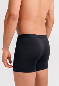 Schwarze Boxer-Briefs mit glatter Textur, elastischem Bund mit dezenter Markenkennzeichnung und einem taillierten Schnitt, der sich dem Körper anpasst.