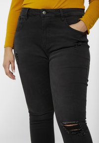 Vaqueros negros de mezclilla con cintura alta, con cierre de botones, bolsillos laterales y rotos desgastados en los muslos. Textura suave, ajuste slim.