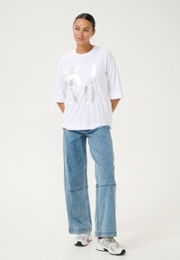 T-shirt blanc avec l'inscription en lettres argentées et audacieuses "KIND", associé à un jean en denim bleu ample et des baskets grises. Design simple et décontracté.