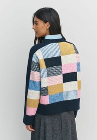 Kobieta o ciemnych włosach, ubrana w kolorowy sweter patchworkowy z niebieskimi, różowymi, żółtymi i białymi wzorami, w połączeniu z ciemną spódnicą.