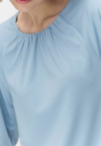 Lyseblå bluse med samlet halsudskæring og lange ærmer. Stoffet virker glat med en blød tekstur og en let glans.