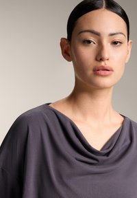 Labava, drapirana majica od tamno sive tkanine. Ima cowl izrez i mekani tekstur, široke otvorene rukave i minimalne detalje.