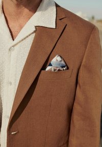 Brauner Blazer mit strukturierter Oberfläche, kombiniert mit einem weißen Hemd mit geometrischem Muster und einem gefalteten Taschentuch in Blau, Pink und Weiß.