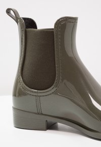 Bota de lluvia de tobillo en color verde oliva brillante con panel lateral elástico y tirador, con tacón bajo apilado sobre fondo blanco.